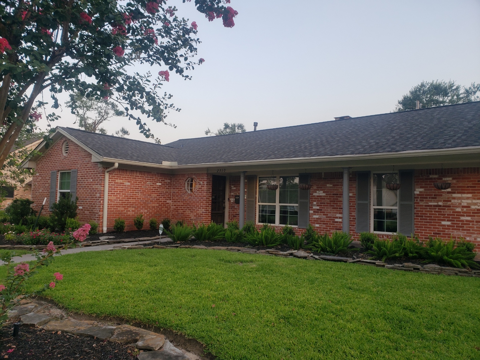 Stylish residential home exterior with red brick façade, new roofing, landscaped garden, and vibrant flowers, showcasing Pride Construction & Roofing's remodeling expertise.