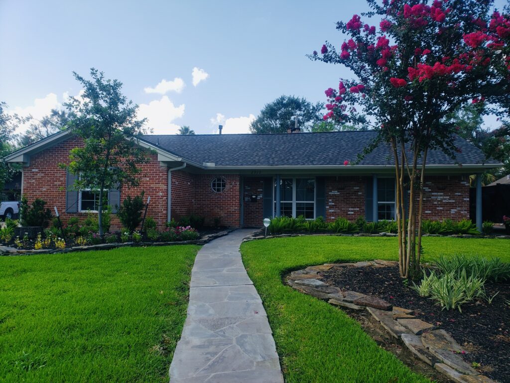 Residential home in Houston Heights with new high-performance shingles, landscaped yard featuring flowers and shrubs, and a stone walkway leading to the entrance.