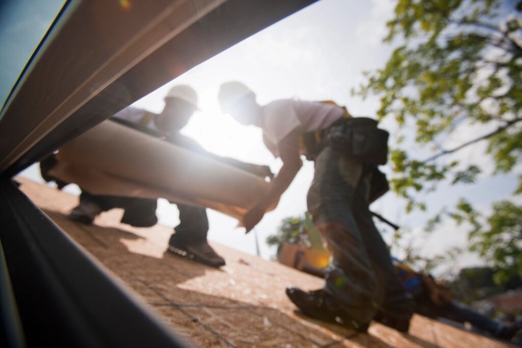 Roofers working on a commercial roofing project, handling materials and tools, with sunlight and trees in the background, representing Pride Construction & Roofing's services in Houston.