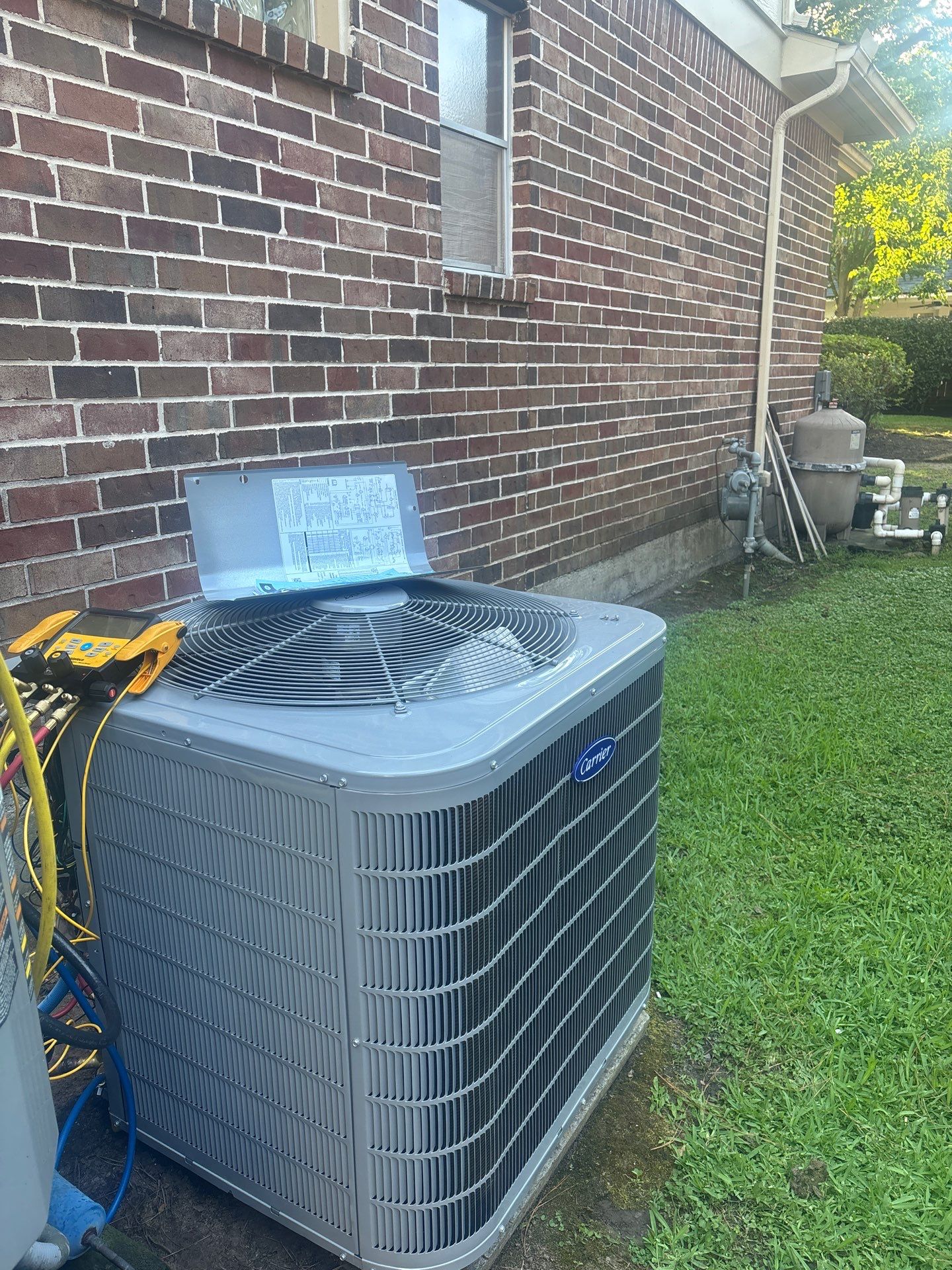 Air conditioning unit installed beside a brick house, featuring visible service gauges and a well-maintained lawn, illustrating home comfort solutions by Pride Construction & Roofing.