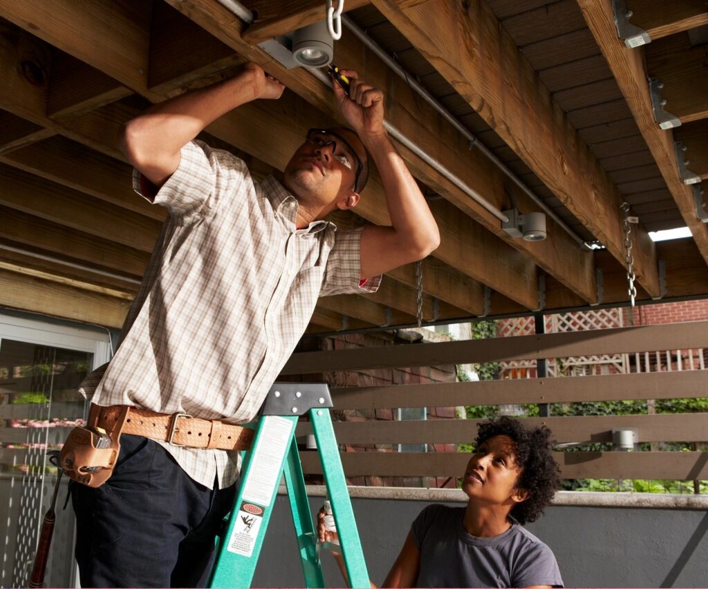 Person on ladder performing home maintenance, inspecting light fixture, and assisting partner, emphasizing proactive home care services by Pride Construction & Roofing.