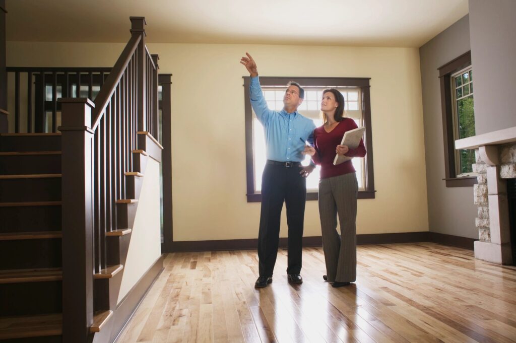 Man pointing at ceiling while discussing home improvements with a woman holding a clipboard in a well-lit interior space.