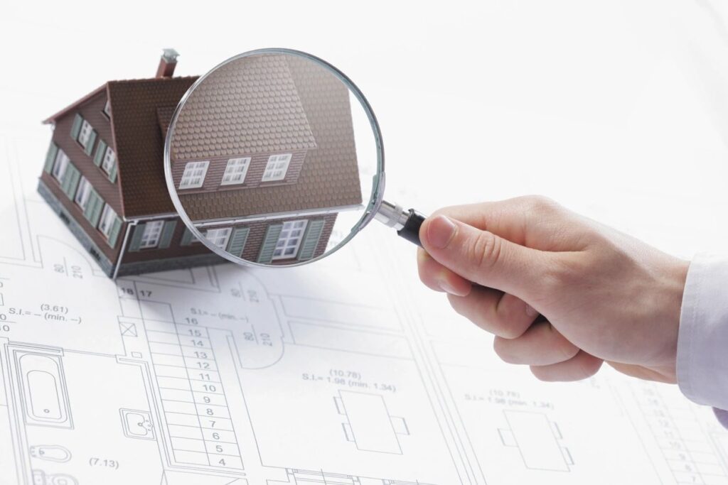 Hand holding a magnifying glass over a model house on architectural blueprints, symbolizing detailed home inspections and maintenance for proactive home care.